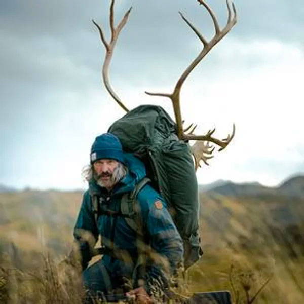 Man hiking through grassy hills with a large pack carrying antlers strapped on top under an overcast sky.