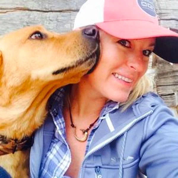 Woman in a baseball cap smiles as a golden-brown dog nuzzles her cheek outdoors in front of a wooden wall.