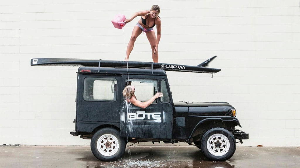A woman pours water from a jerry can onto a friend climbing out of a Jeep with a paddleboard strapped to the roof.