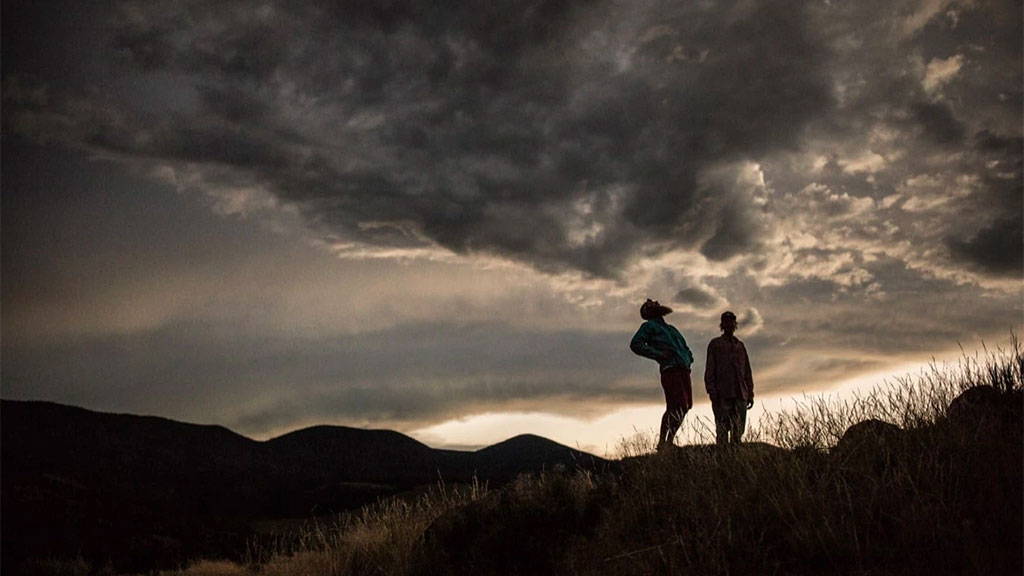 Two people standing on a grassy ridge at dusk beneath dramatic storm clouds over distant mountains.