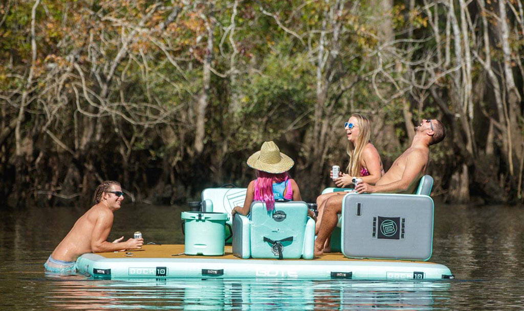 Group of friends relaxing on a floating lounge platform in a wooded river, laughing and holding drinks.