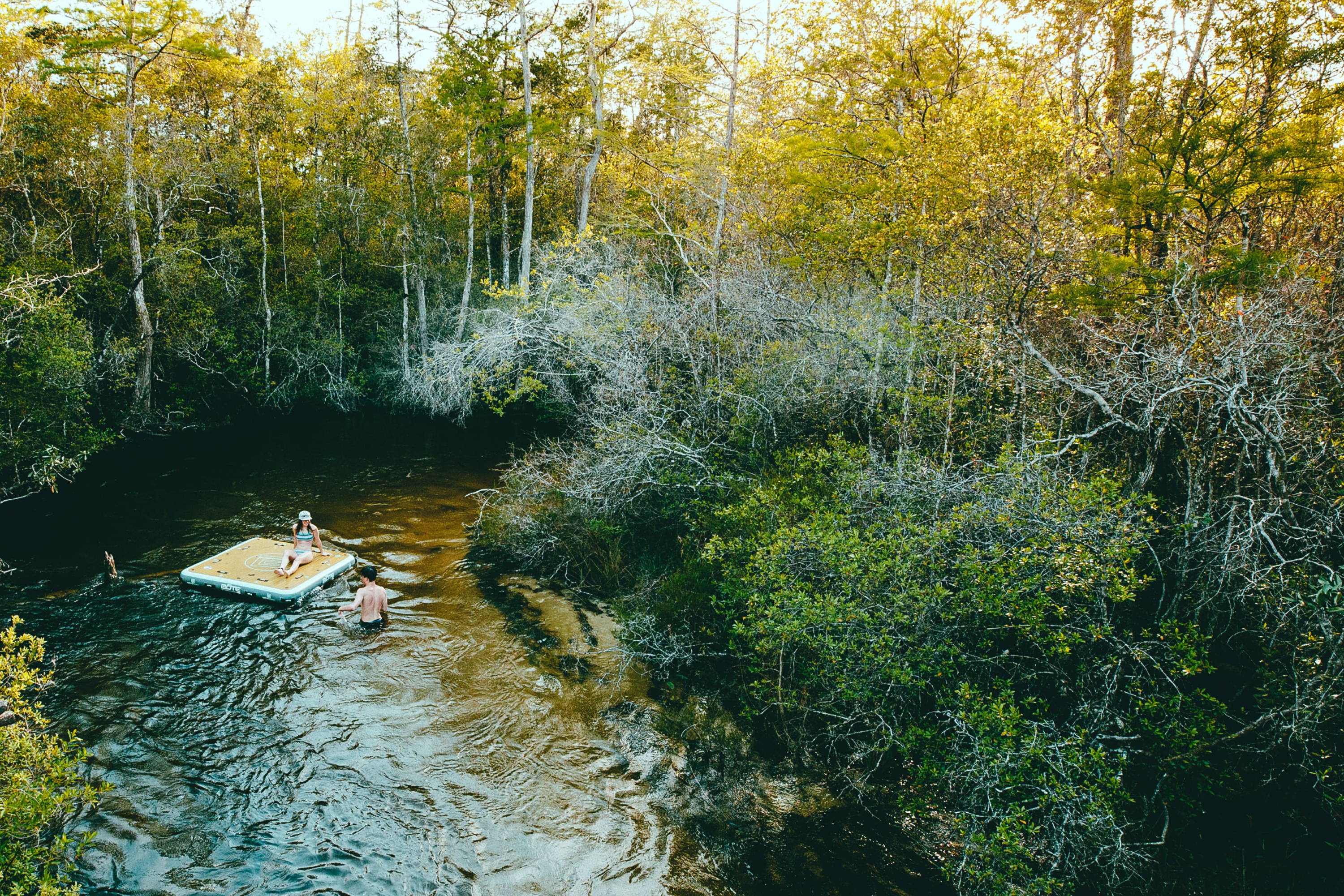 Two people relaxing on a floating water platform in a calm forest creek surrounded by autumn trees.