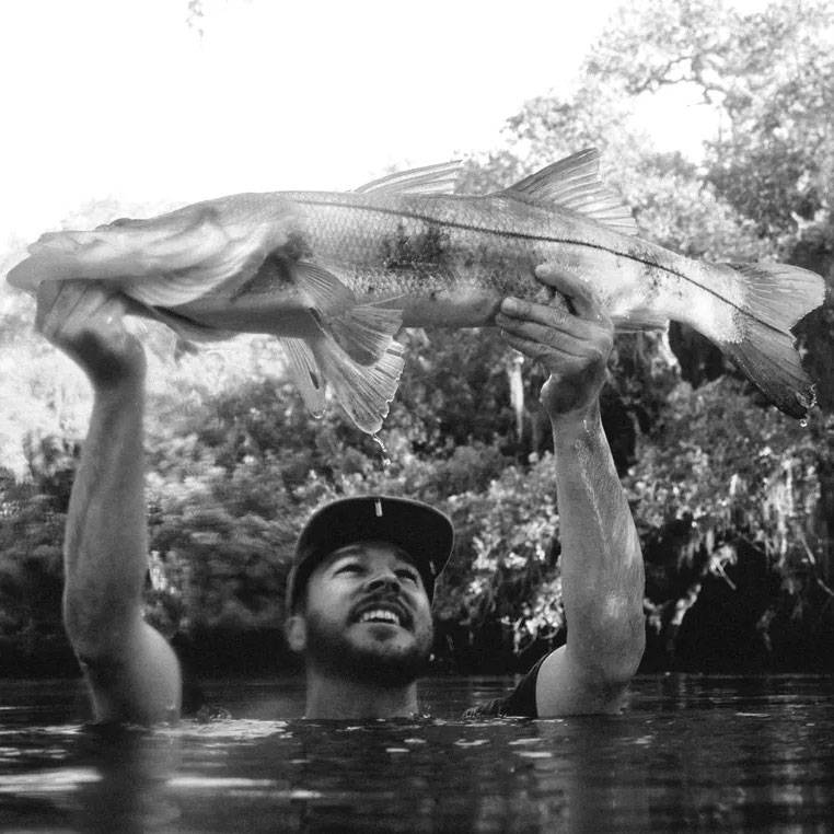 A man standing chest-deep in water lifts a large fish above his head, smiling as trees line the riverbank behind him.