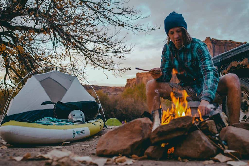 Person tending a campfire beside a tent and inflatable paddle board at a desert campsite.