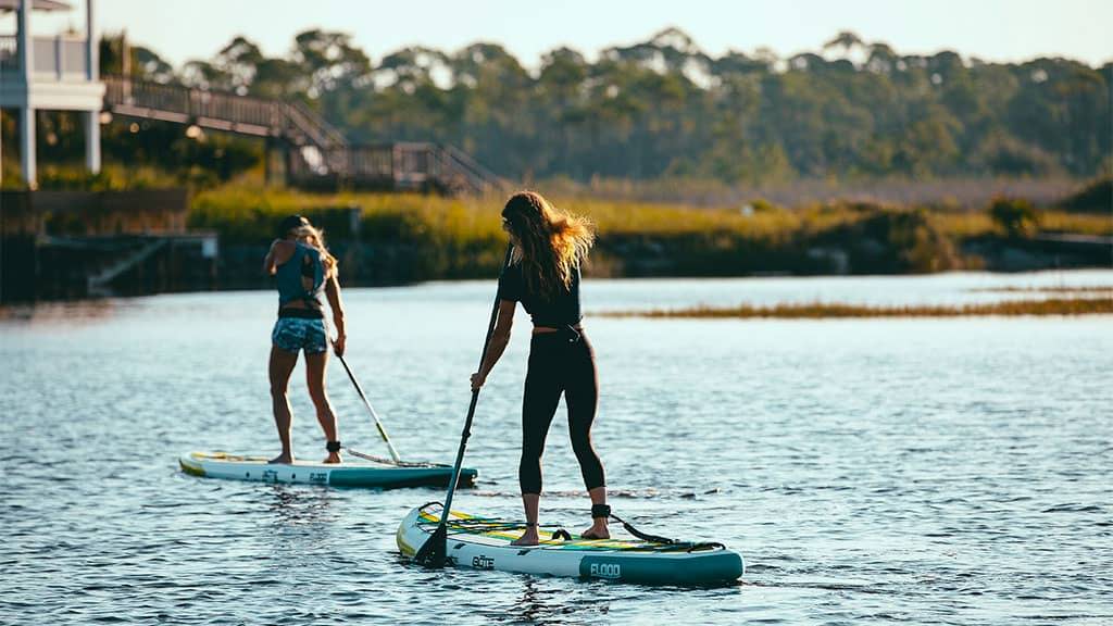 Two people paddle boarding on a calm river near a marshy shoreline with a boardwalk in the background.
