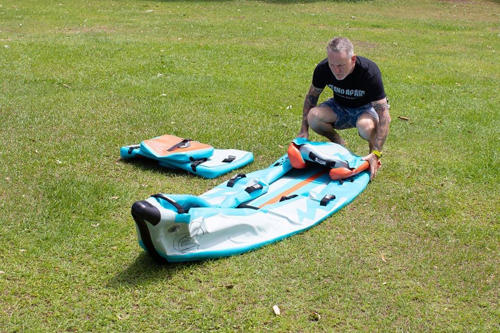Man kneeling on grass rolling up a teal inflatable kayak with orange deck pad and black straps.