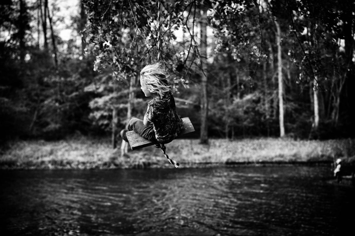 Black and white photo of a child sitting on a rope swing over a quiet forest river.