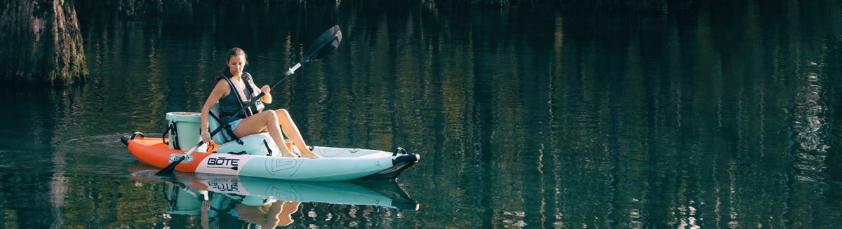 Woman paddling inflatable kayak on calm water with trees reflecting around her.