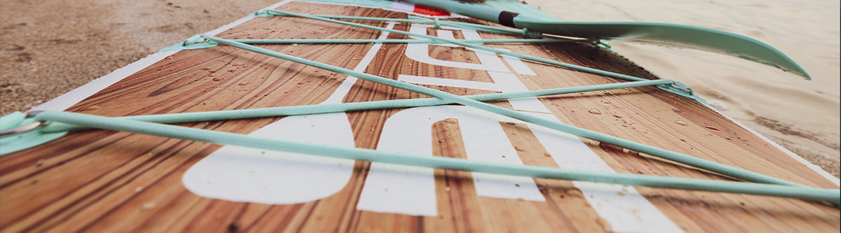 Close-up of woodgrain paddle board with mint bungee cords resting on the sand.