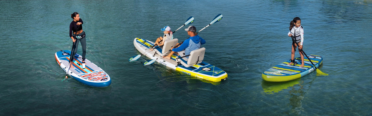 Group paddling on calm blue water, including adults and child on paddle boards and kayak-style board.