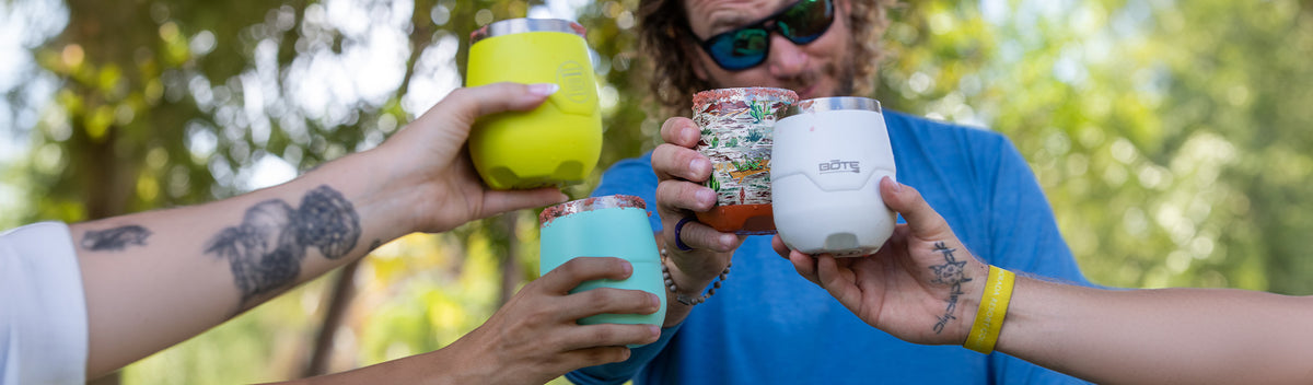 Close-up of four people toasting colorful stemless cups outdoors with blurred greenery in background.