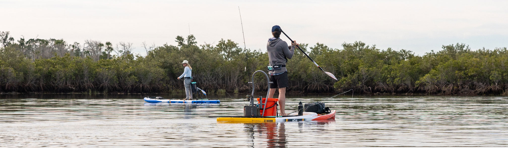 Woman fishing from blue inflatable paddle board near trees on calm water.