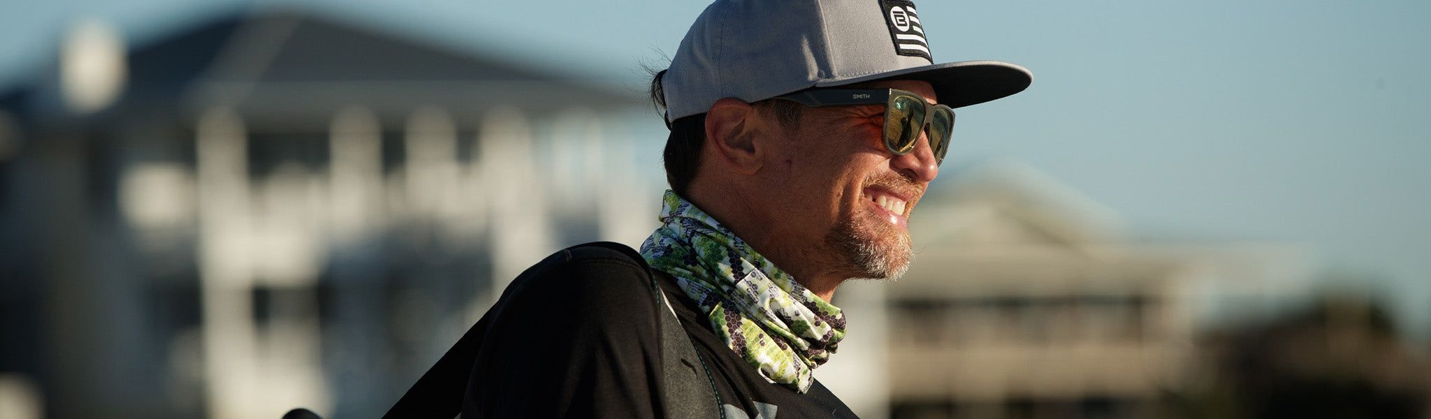 Smiling man wearing gray hat and camo neck gaiter outdoors on a sunny day.