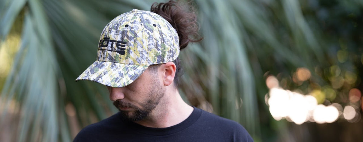 Man wearing a camo hat with logo, looking down against blurred palm leaves.