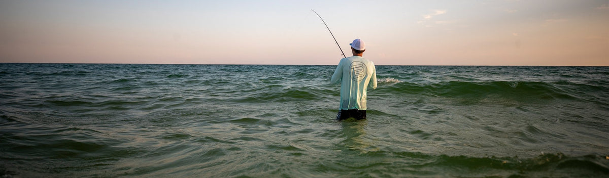 Angler standing waist-deep in ocean waves fishing at sunset under a soft pastel sky.
