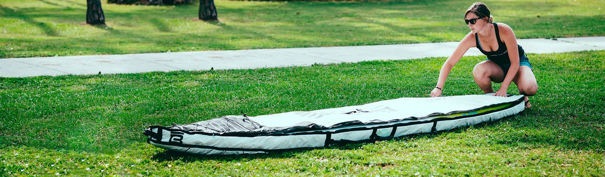 Woman zipping paddle board bag on grass near a sidewalk in bright sunlight.