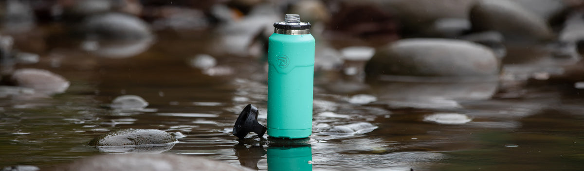 Close-up of turquoise MAGNEBottle standing in shallow stream surrounded by smooth rocks.