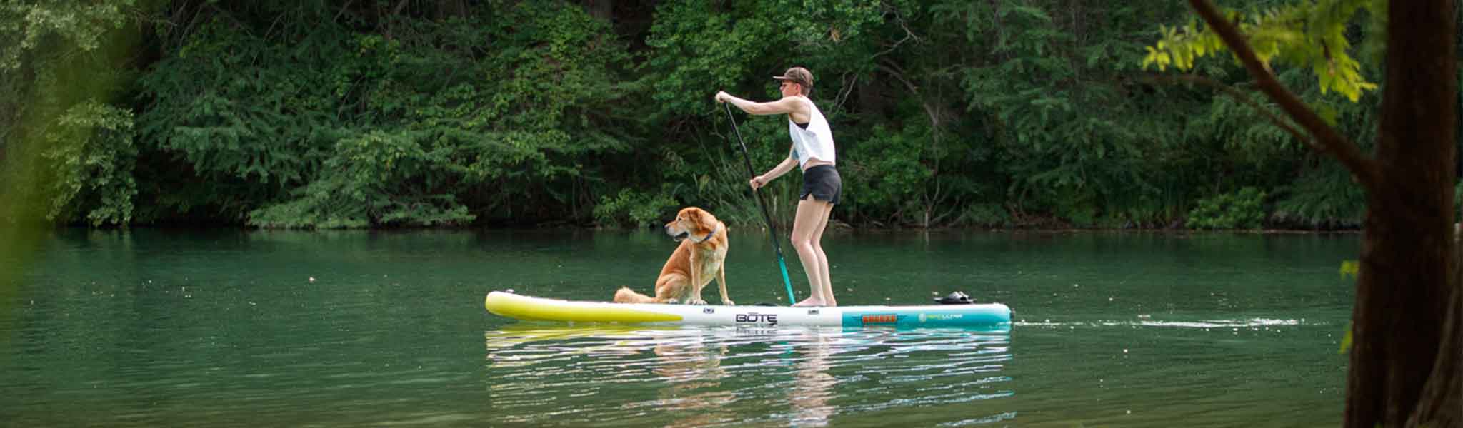 Woman paddling with dog on board in calm green water surrounded by trees.