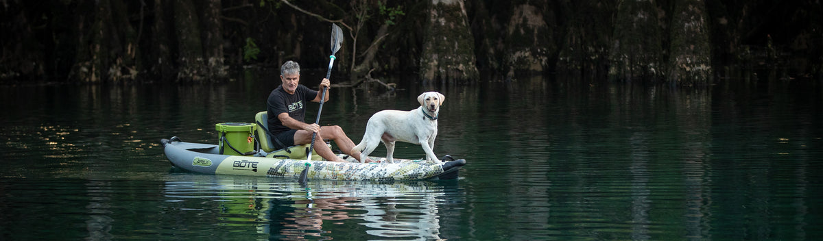 Man paddling inflatable kayak with dog standing on front deck in calm wooded water.