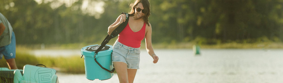 Woman carrying teal cooler with shoulder strap along lake shore in sunlight.