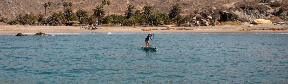 Person paddling near rocky shoreline with palm trees and sandy beach in the background.