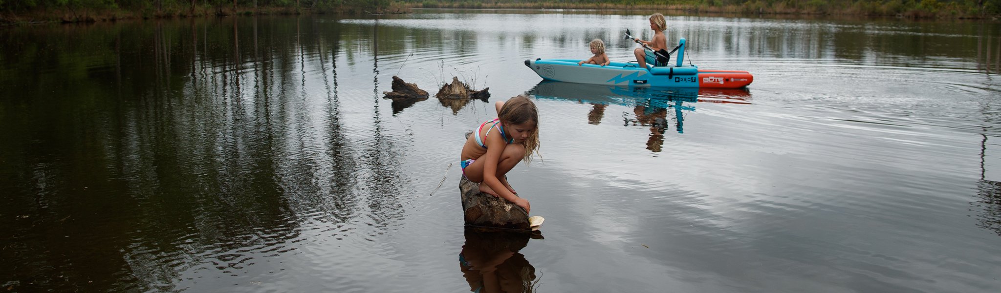 Children playing by a calm lake, one paddling a blue kayak near the shore.