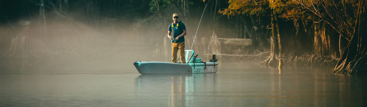 Man fishing from kayak on misty water surrounded by trees at sunrise.