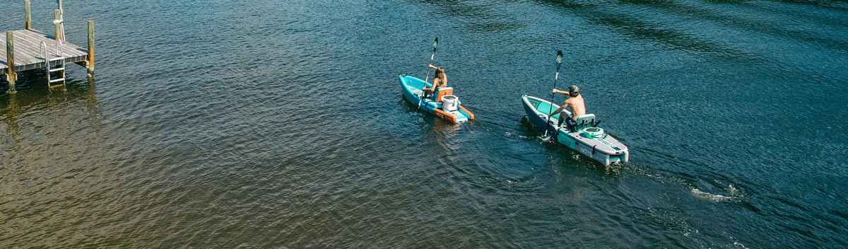 Aerial view of two people paddling kayaks near a wooden dock on open water.