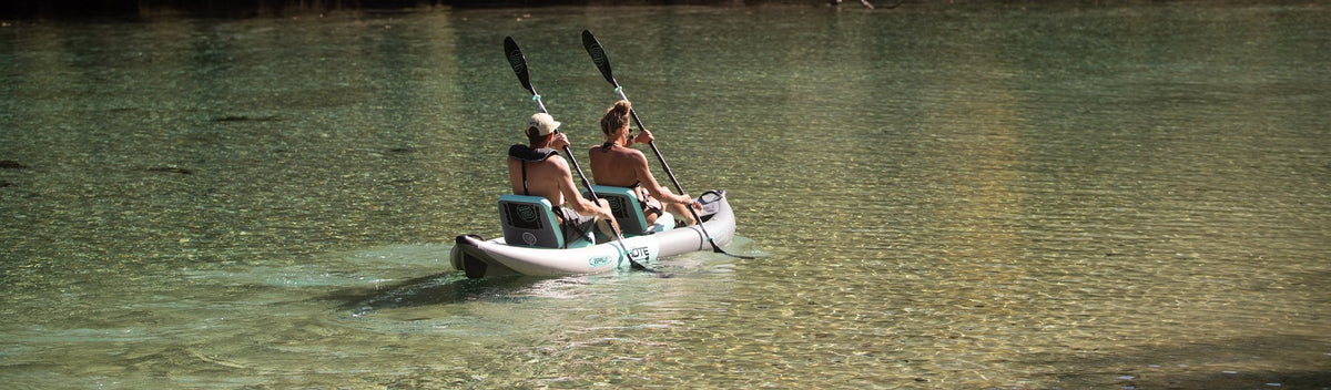 Two people paddling tandem inflatable kayak across clear shallow water.