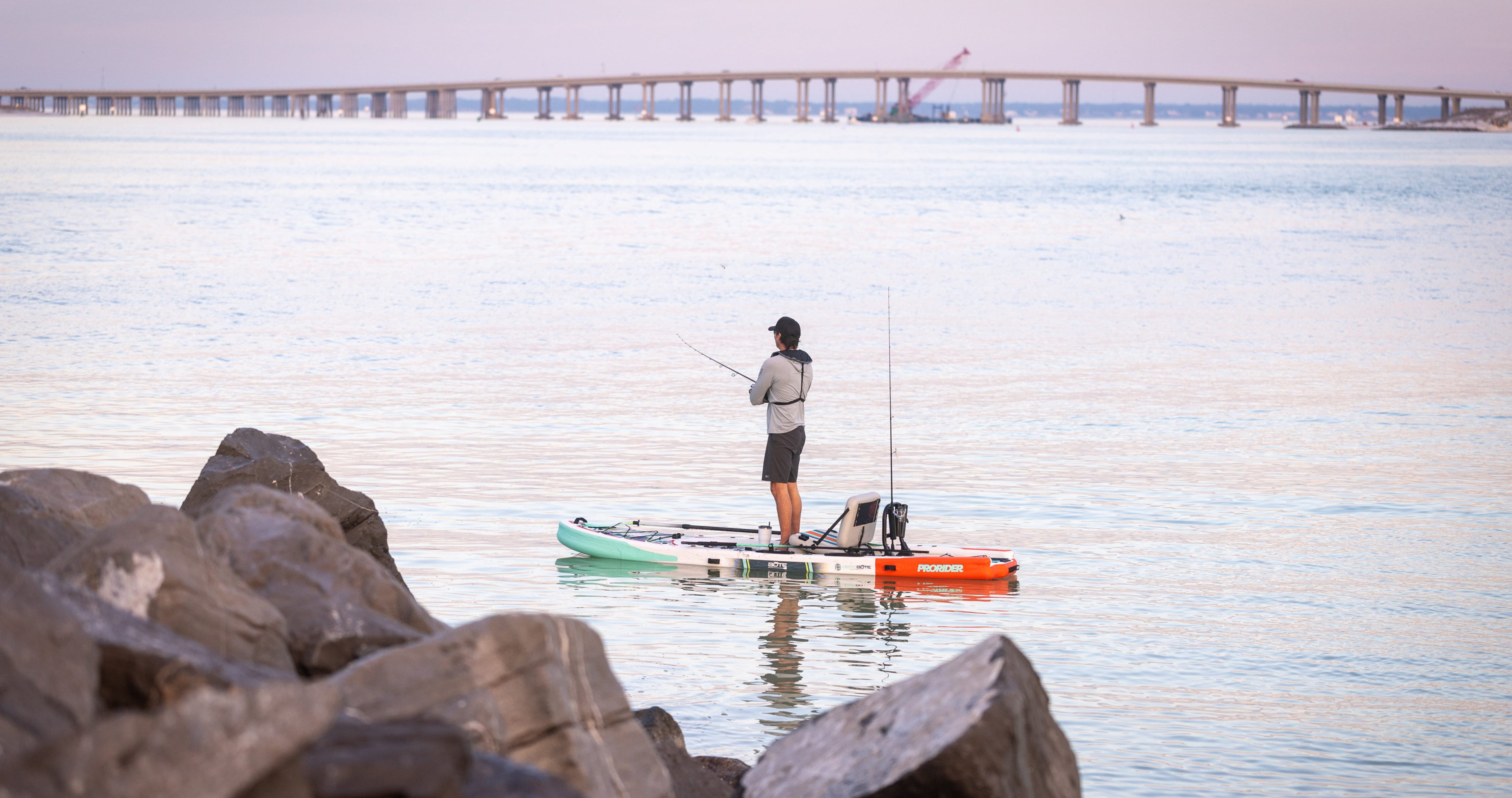 Person fishing from a stand-up paddleboard on a calm body of water with rocks and a bridge in the background.