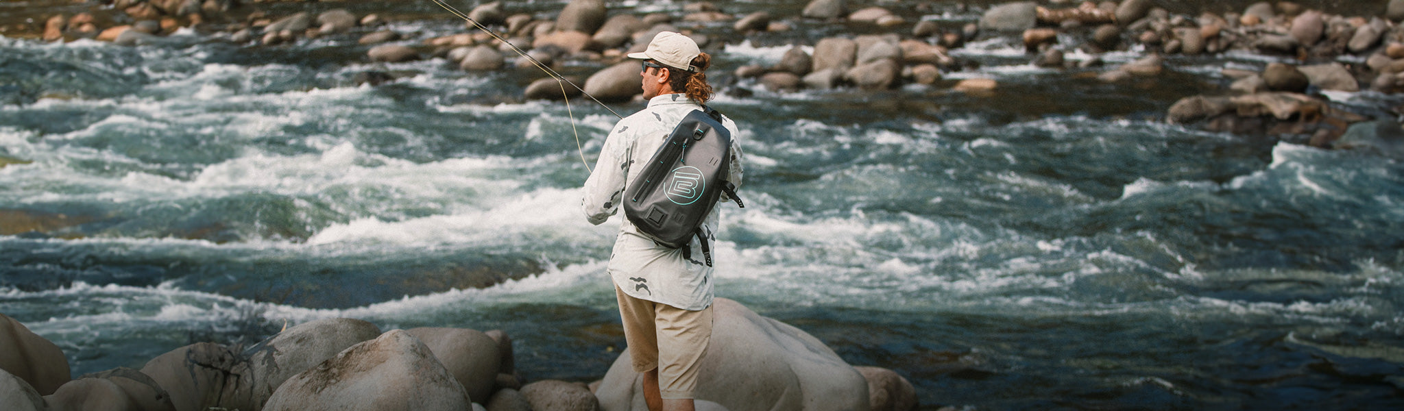 Man fishing beside a rocky river wearing a gray shirt and black waterproof backpack.