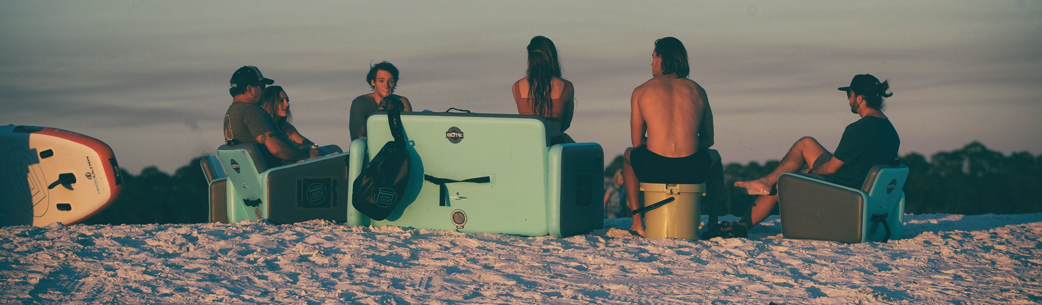 Group sitting on coolers and chairs in the sand during sunset, paddle boards nearby.