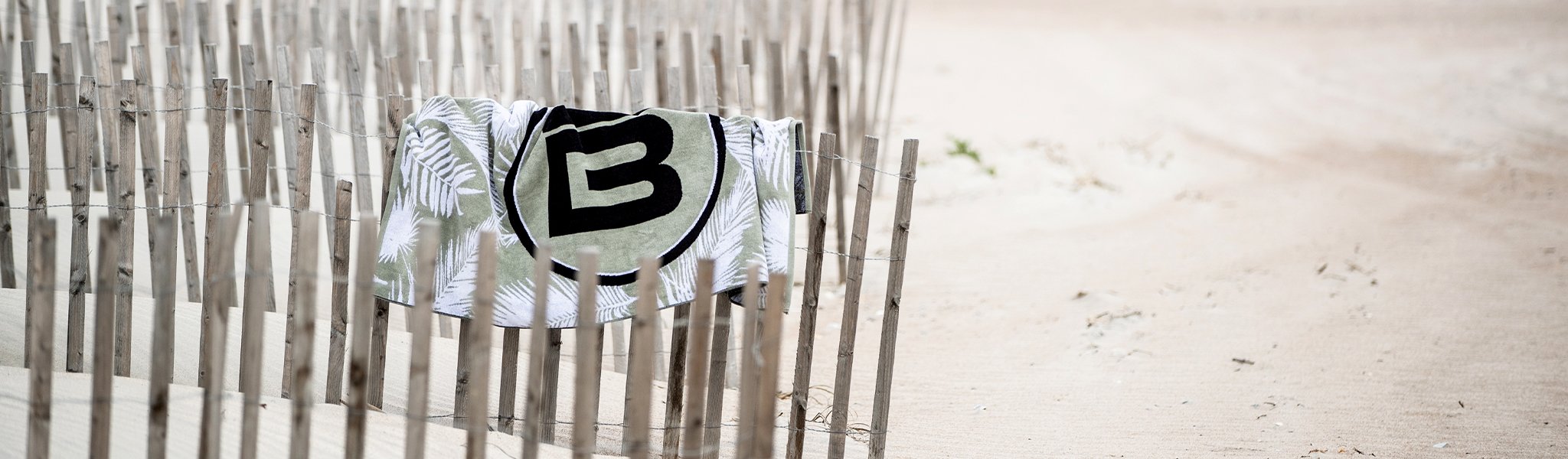 Towel with large logo draped across wooden beach fence in sand dunes.