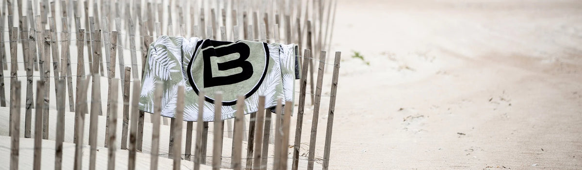 Towel with large logo draped across wooden beach fence in sand dunes.
