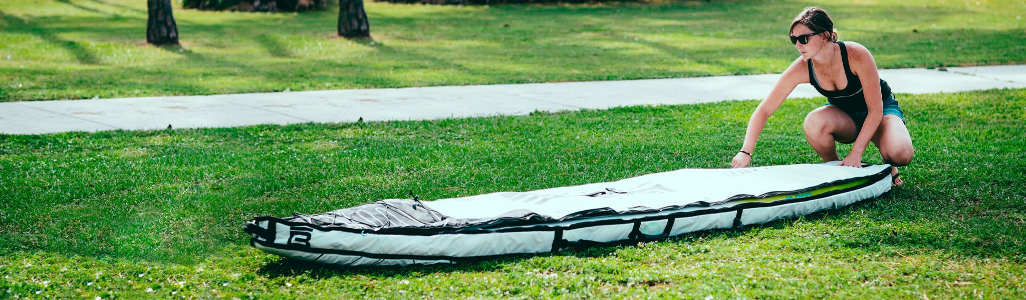 Woman zipping paddle board bag on grass near a sidewalk in bright sunlight.