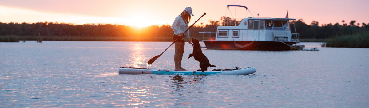 Woman paddle boarding with a black dog at sunset, boat anchored in background.