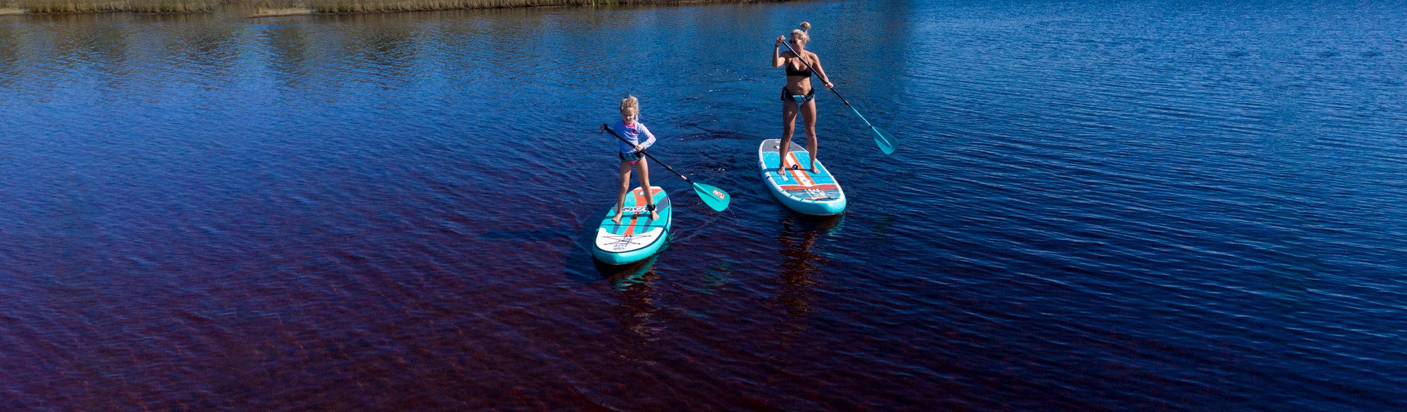 Woman and child paddle boarding together on calm blue water under clear skies.