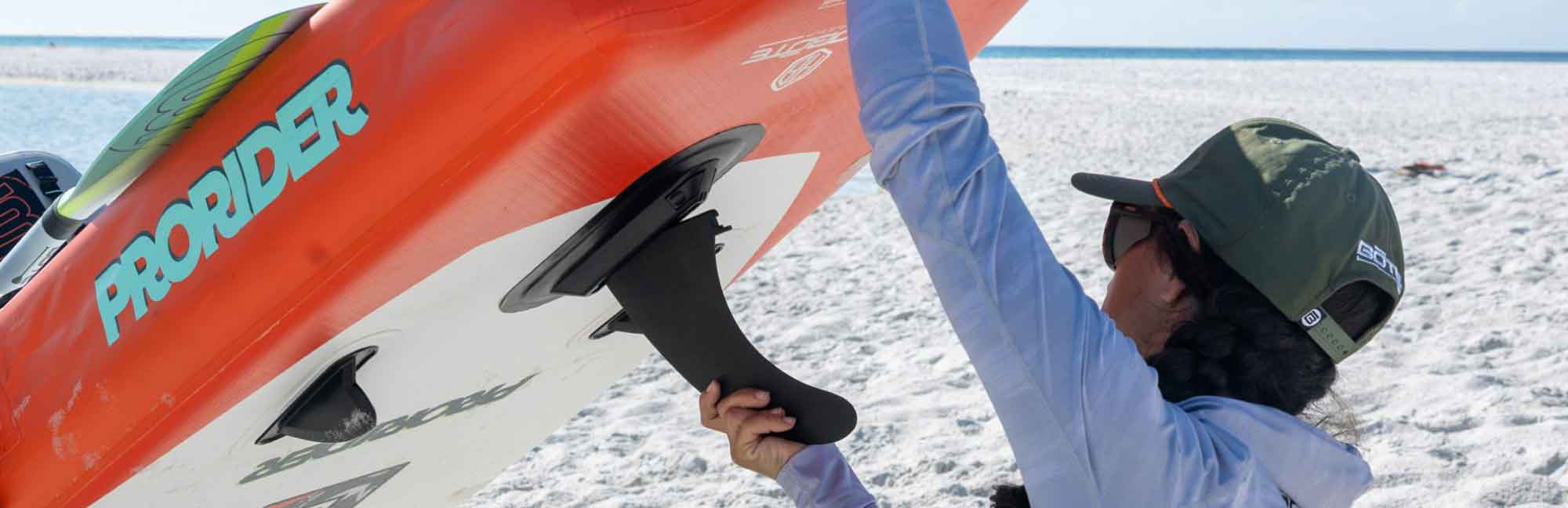 Person holding a black fin attached to an orange paddle board with 'ProRider' branding on a sandy beach.