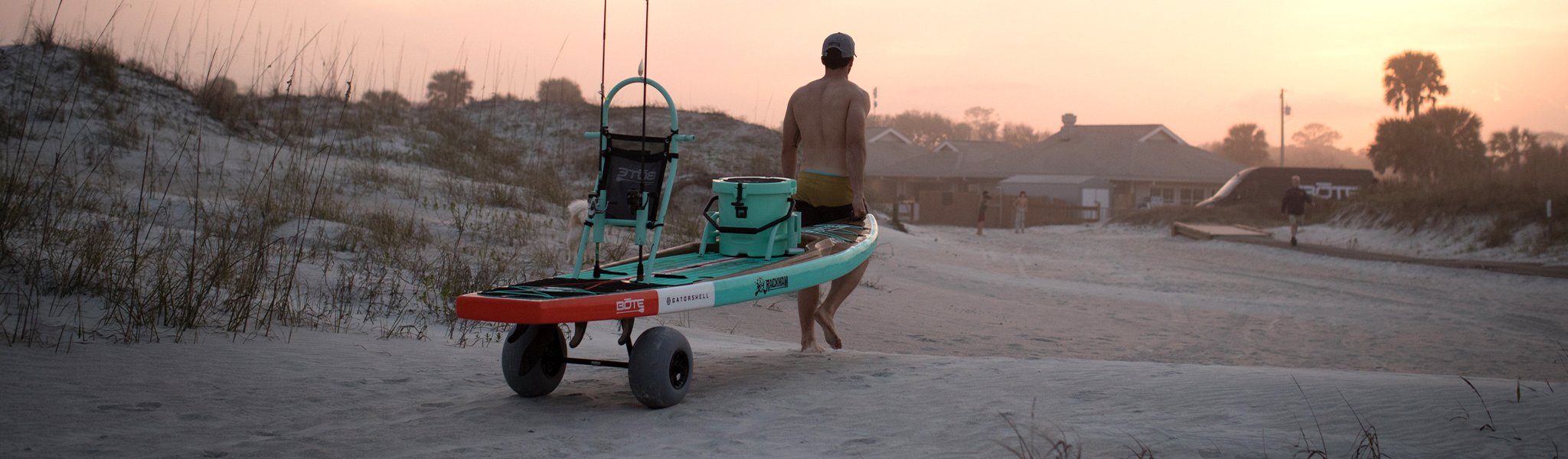 Man pulling fishing paddle board on sand at sunset with dunes in background.