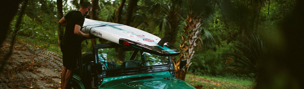 Man securing paddle board to the roof of a green jeep surrounded by palm trees.