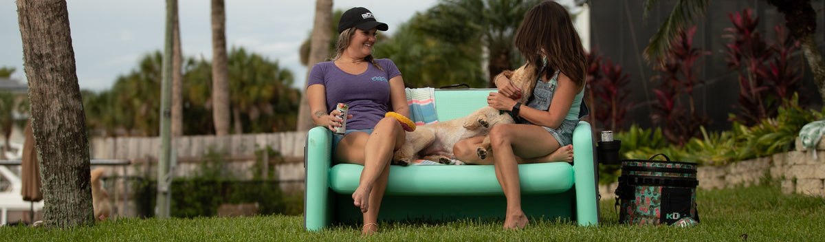 Two women sitting on inflatable couch with dog and drinks in a backyard setting.