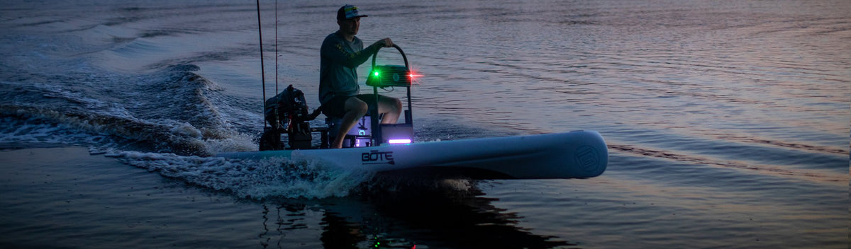 Man driving small powered skiff across calm water at dusk with navigation lights on.