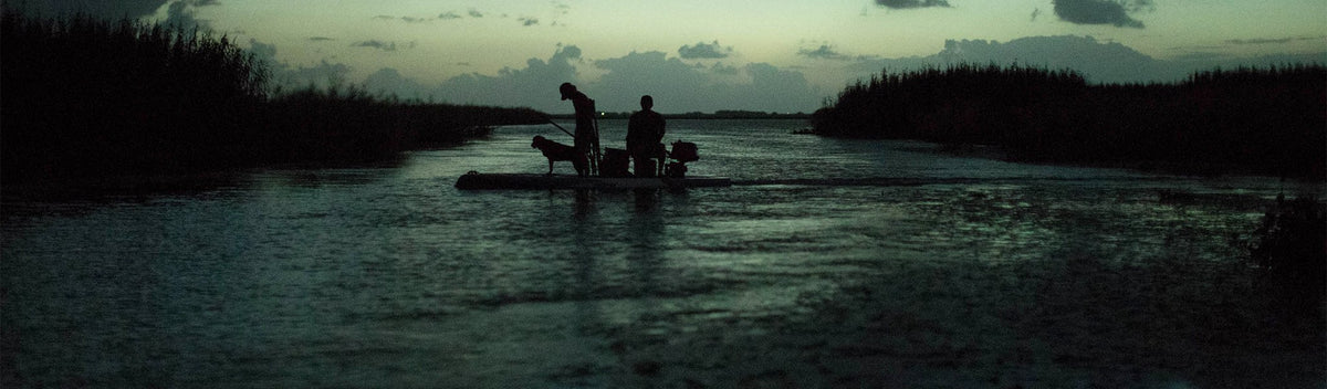 Two people and a dog riding motorized skiff through narrow waterway at dusk.