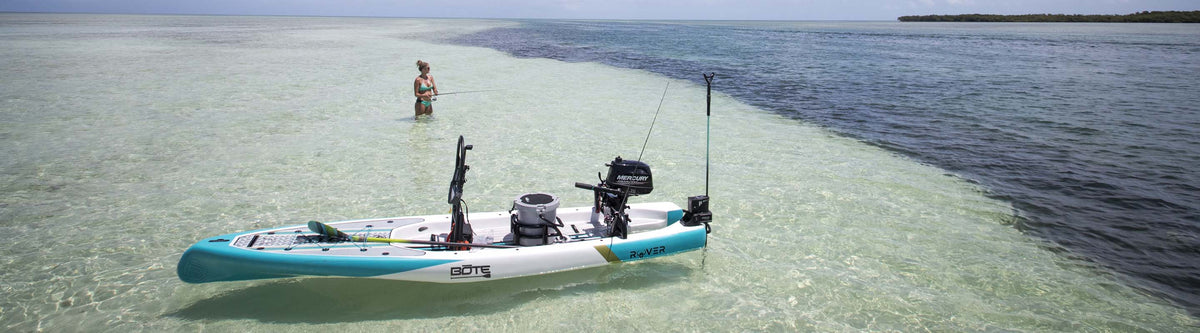 Woman fishing in shallow clear water beside motorized paddle board loaded with gear.