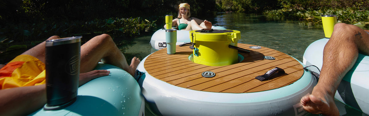 People relaxing on inflatable floats with round table holding cups and cooler on the water.