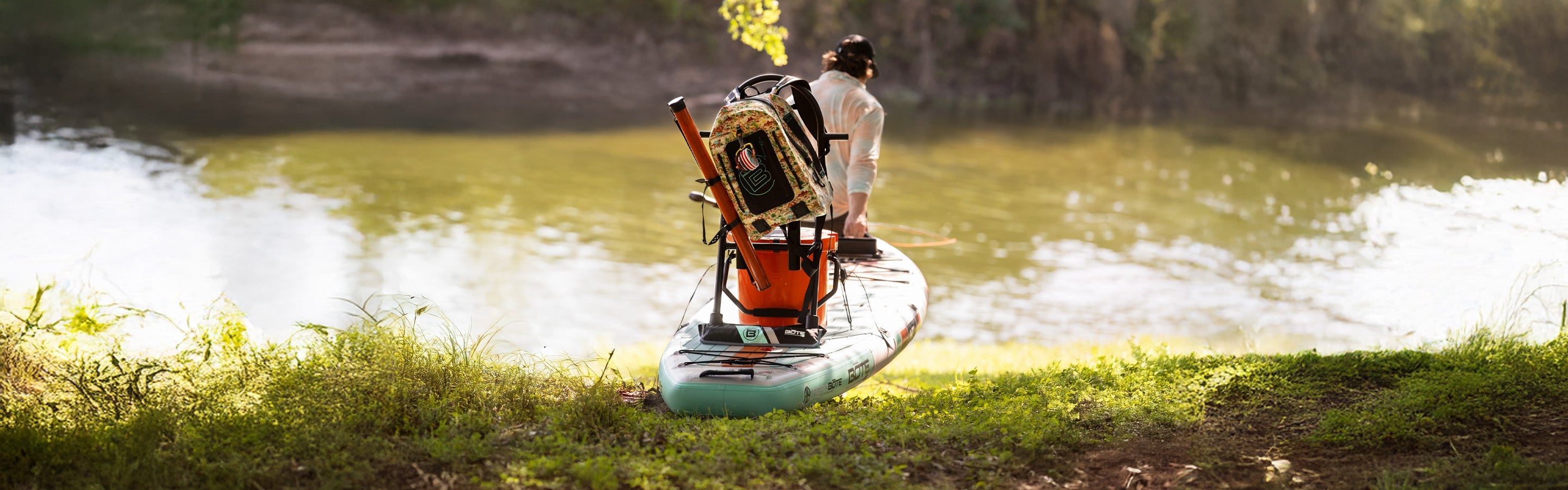 Angled view of paddle board on grassy shore with mounted gear and paddler standing by the water.