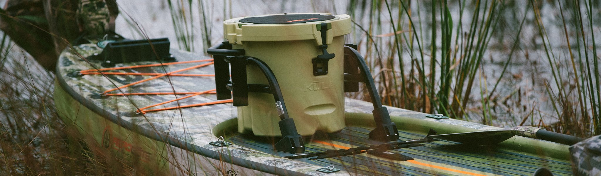 Close-up of tan KULA cooler secured on camo paddle board with paddle and grass in background.
