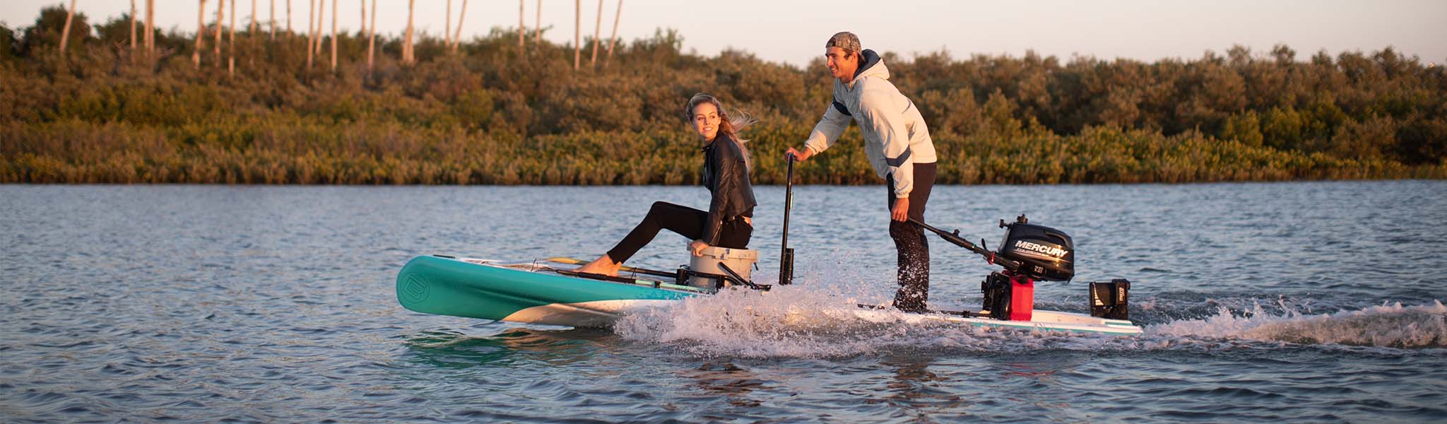 Man and woman riding motorized micro skiff across calm water at sunset.