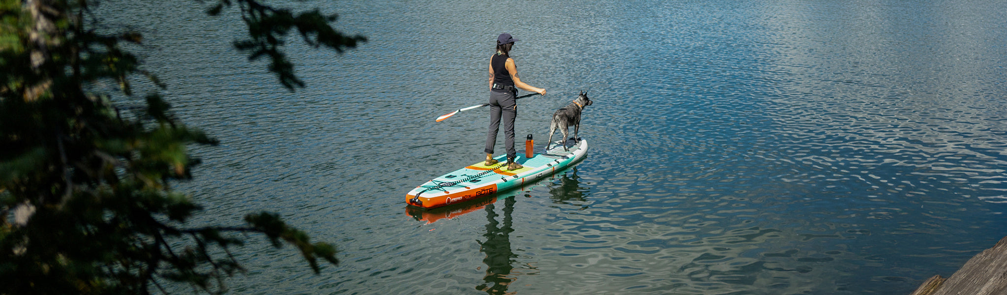 Woman paddle boarding with dog on turquoise lake surrounded by trees.