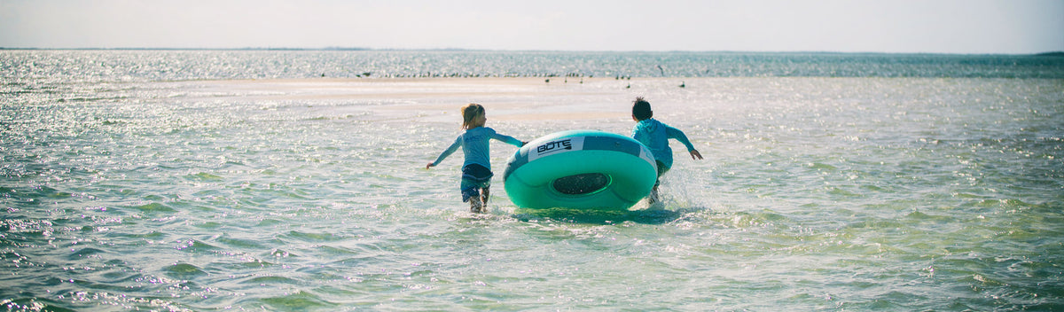 Two kids running into shallow ocean water pulling inflatable tube toward small sandbar.